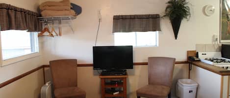 A compact living space with a television, two chairs, a small shelf, a heater, a trash bin, a stove, and a potted plant.
