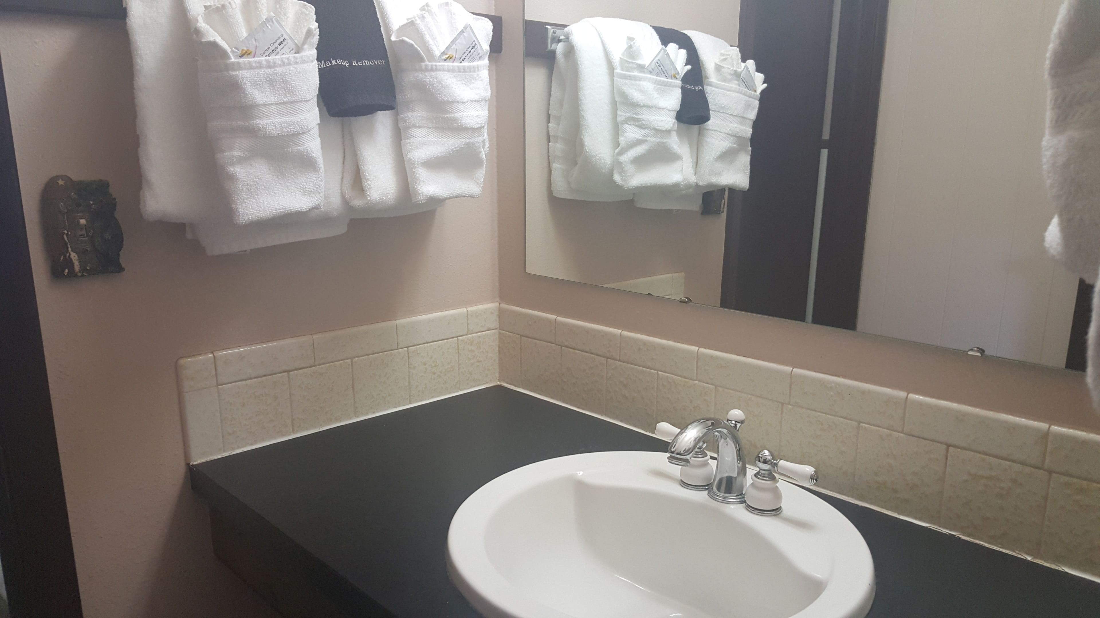 Un baño con un lavamanos blanco, encimera negra y un espejo encima. Hay toallas colgadas de ganchos y un pequeño objeto decorativo en la pared.