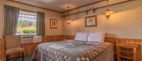 A bedroom with a large bed, a chair, a window with curtains, a wall-mounted lamp, and a framed picture.