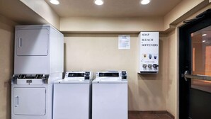 Une buanderie équipée de trois lave-linge et trois sèche-linge, un panneau indiquant les produits de lessive disponibles, et une porte située sur la droite.