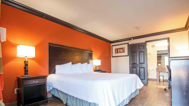 Une chambre d’hôtel avec un grand lit, deux tables de chevet avec des lampes, une tête de lit en bois et une porte donnant sur une autre pièce.