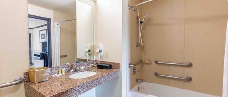 Une salle de bain dotée d’un lavabo avec un plan de travail en granit, d’un miroir, d’une douchette à main et d’une baignoire équipée de barres d’appui.