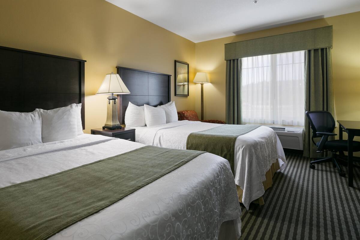 Une chambre d’hôtel avec deux lits, un bureau, une chaise, une lampe et une fenêtre avec des rideaux.