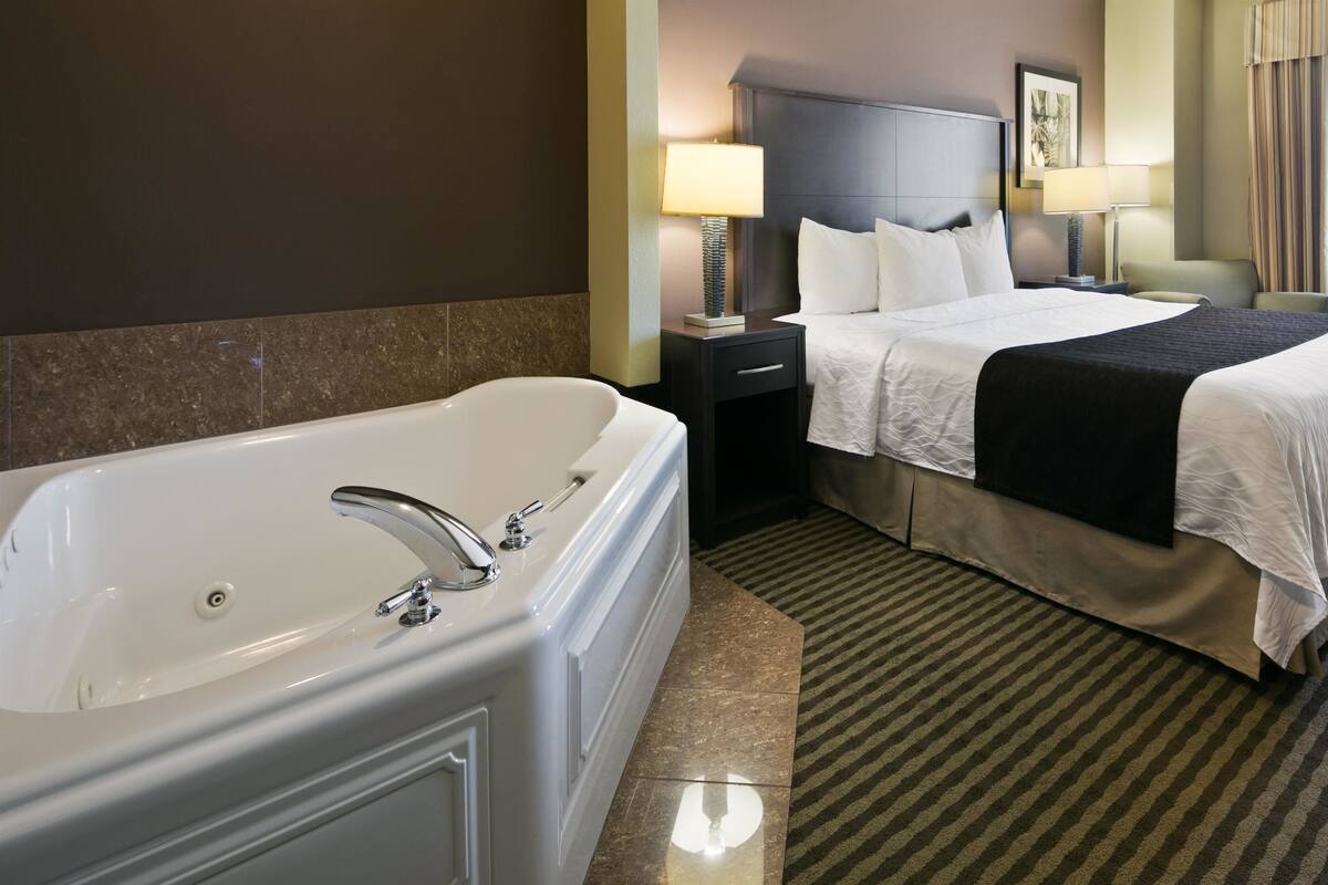 Une chambre d’hôtel avec une grande baignoire blanche, deux lits avec du linge de lit blanc, une tête de lit sombre, une petite table de chevet avec une lampe et une fenêtre avec des rideaux.