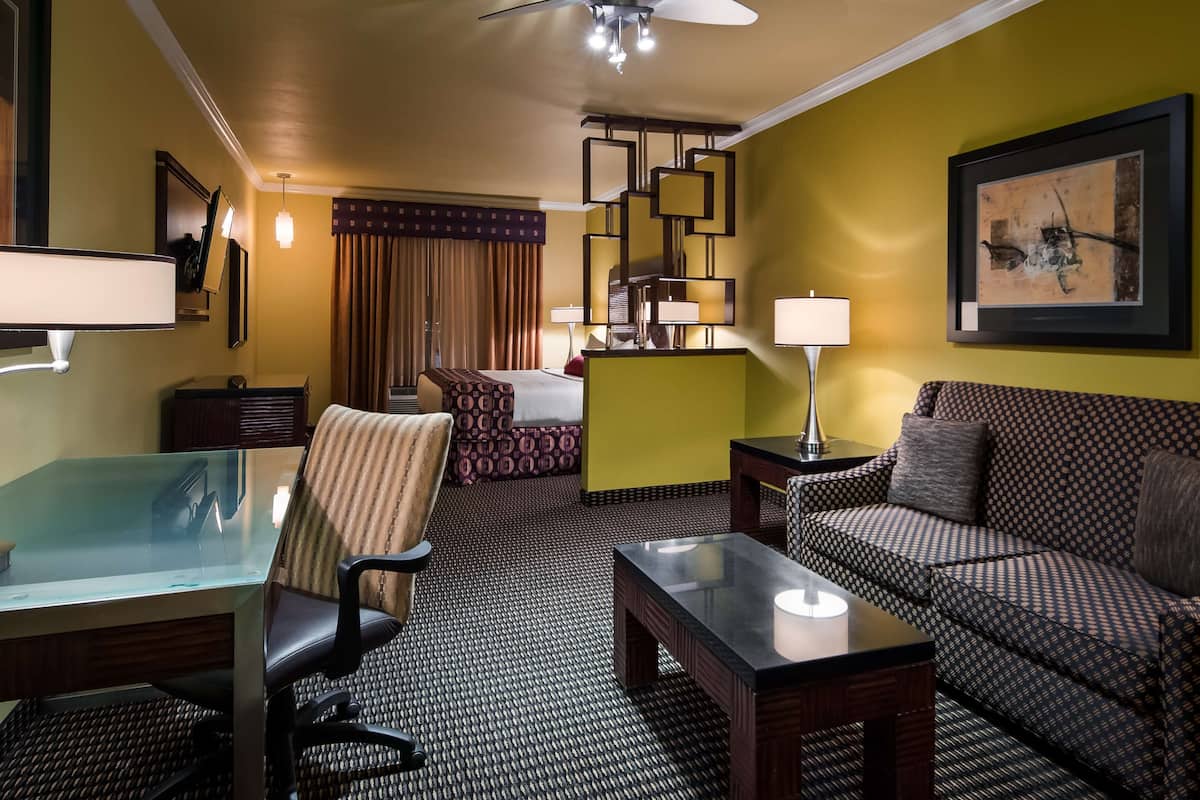 Quarto de hotel com cama, sofá, escrivaninha com cadeira, candeeiro de pé e um quadro na parede.