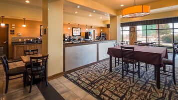 A hotel lobby with a reception desk, a dining area with a table and chairs, and a kitchen in the background.