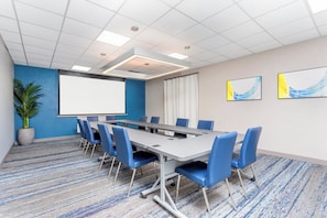 Una sala conferenze con un lungo tavolo, sedie blu, uno schermo per proiettore e una pianta in vaso.