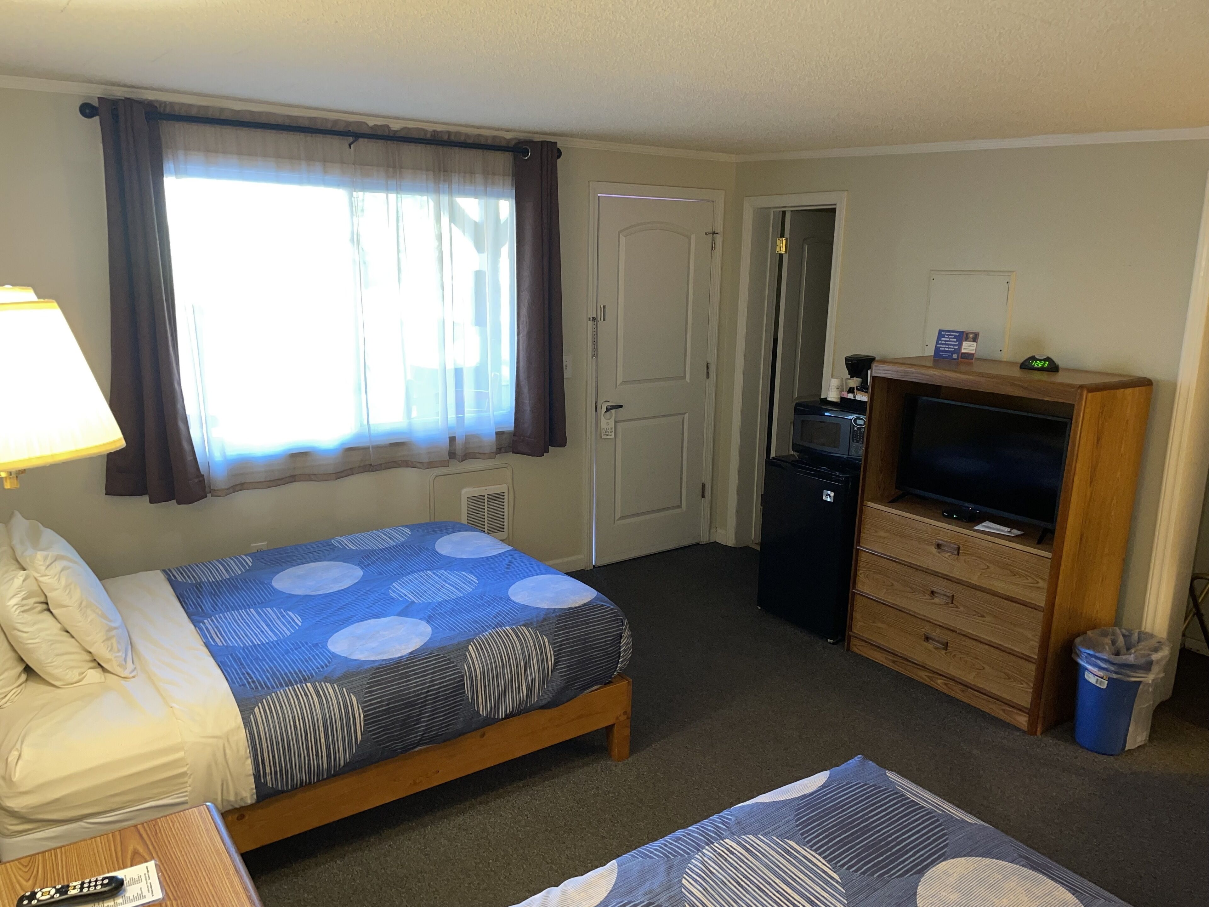 Habitación de hotel con cama, televisión, refrigerador pequeño, cómoda de madera, lámpara y ventana con cortinas.