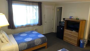 Habitación de hotel con cama, televisión, refrigerador pequeño, cómoda de madera, lámpara y ventana con cortinas.