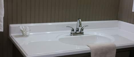 A bathroom with a large white sink, a mirror above it, and a towel rack. There is a small glass container on the sink counter.