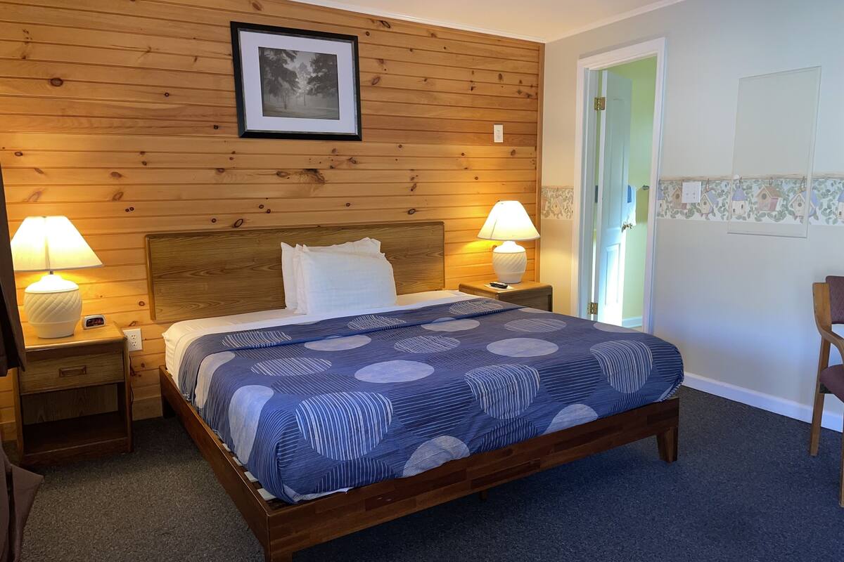 A bedroom with a wooden headboard, a bed with a blue patterned bedspread, two bedside lamps, and a framed picture on the wall.