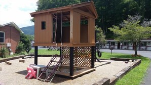 A wooden playhouse with a ladder, a swing set, and a small pink chair in a landscaped area with a picnic table and a tire swing. - A Holiday Motel (Maggie Valley)