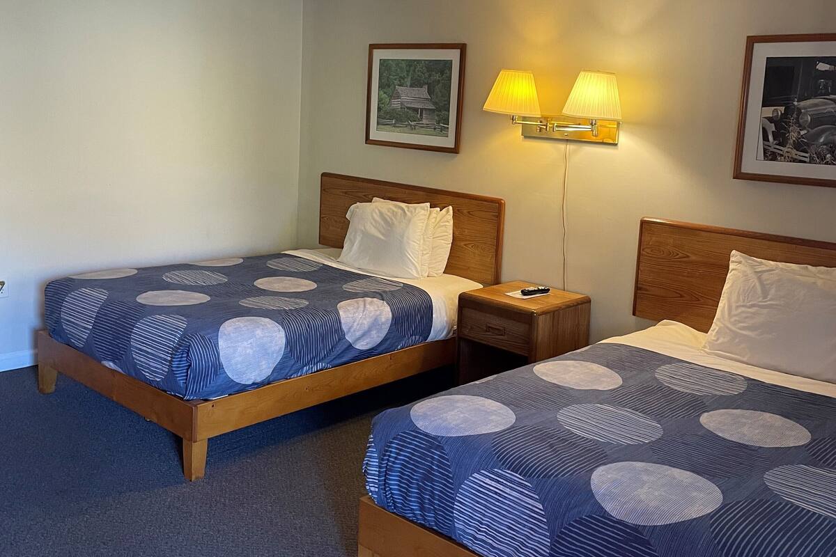 A hotel room with two beds, each with a wooden headboard and a blue patterned bedspread. There are two bedside lamps and a framed picture on the wall.