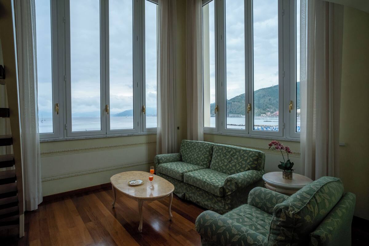 Una habitación con un sofá verde estampado, una pequeña mesa redonda con una bebida en un vaso, y ventanas amplias con vistas a un cuerpo de agua y tierra a lo lejos.