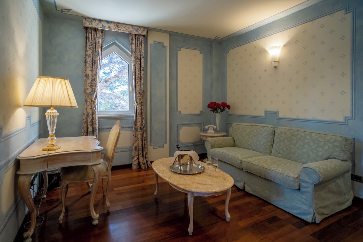 A room with a wooden floor, a light-colored sofa, a small table with a tray, a lamp, a chair, a window with curtains, and a wall-mounted light fixture.