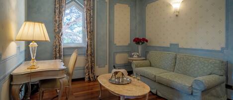 A room with a wooden floor, a light-colored sofa, a small table with a tray, a lamp, a chair, a window with curtains, and a wall-mounted light fixture.