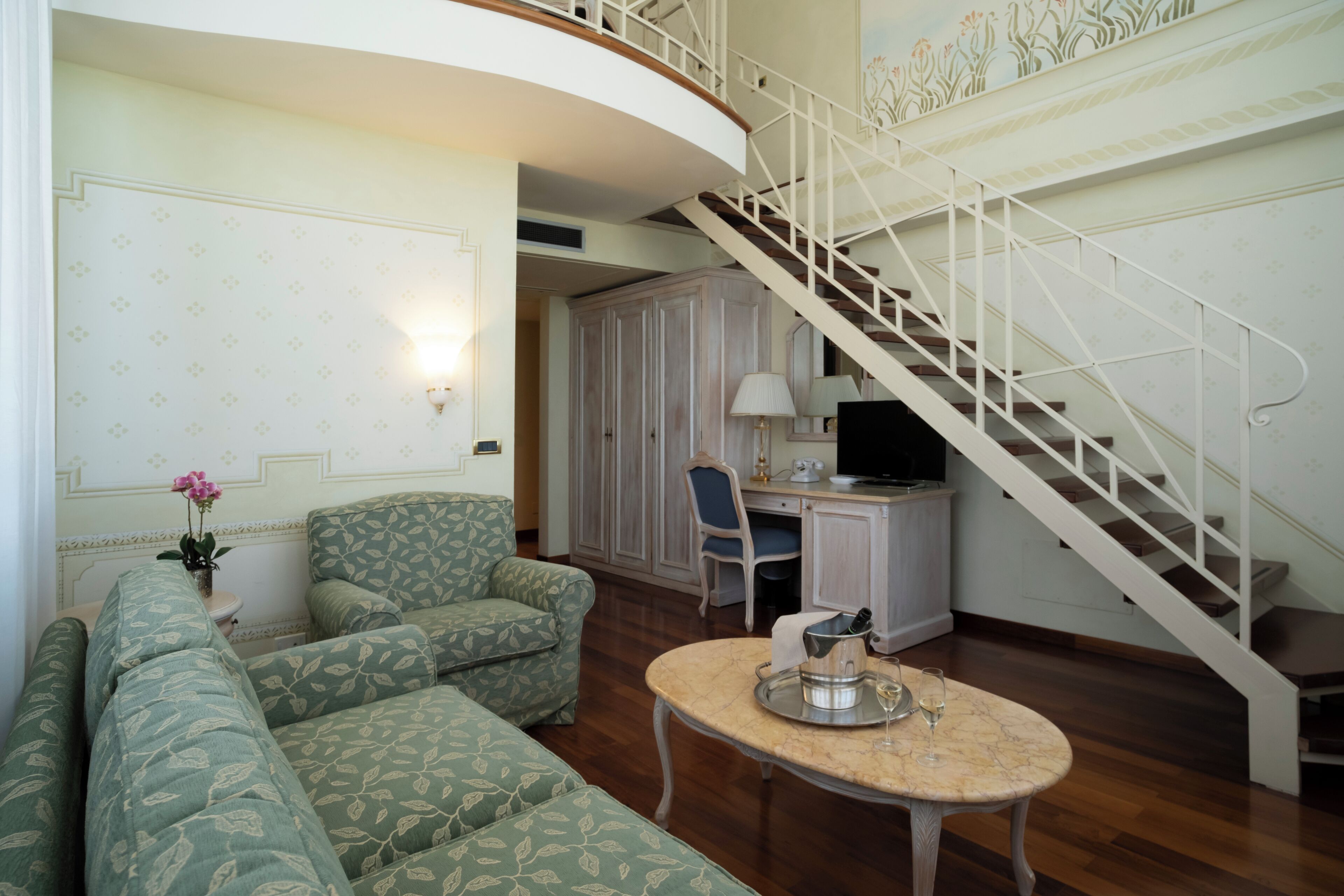 A living room with a curved staircase, green patterned sofas, a round table with a tray, and a desk with a computer.