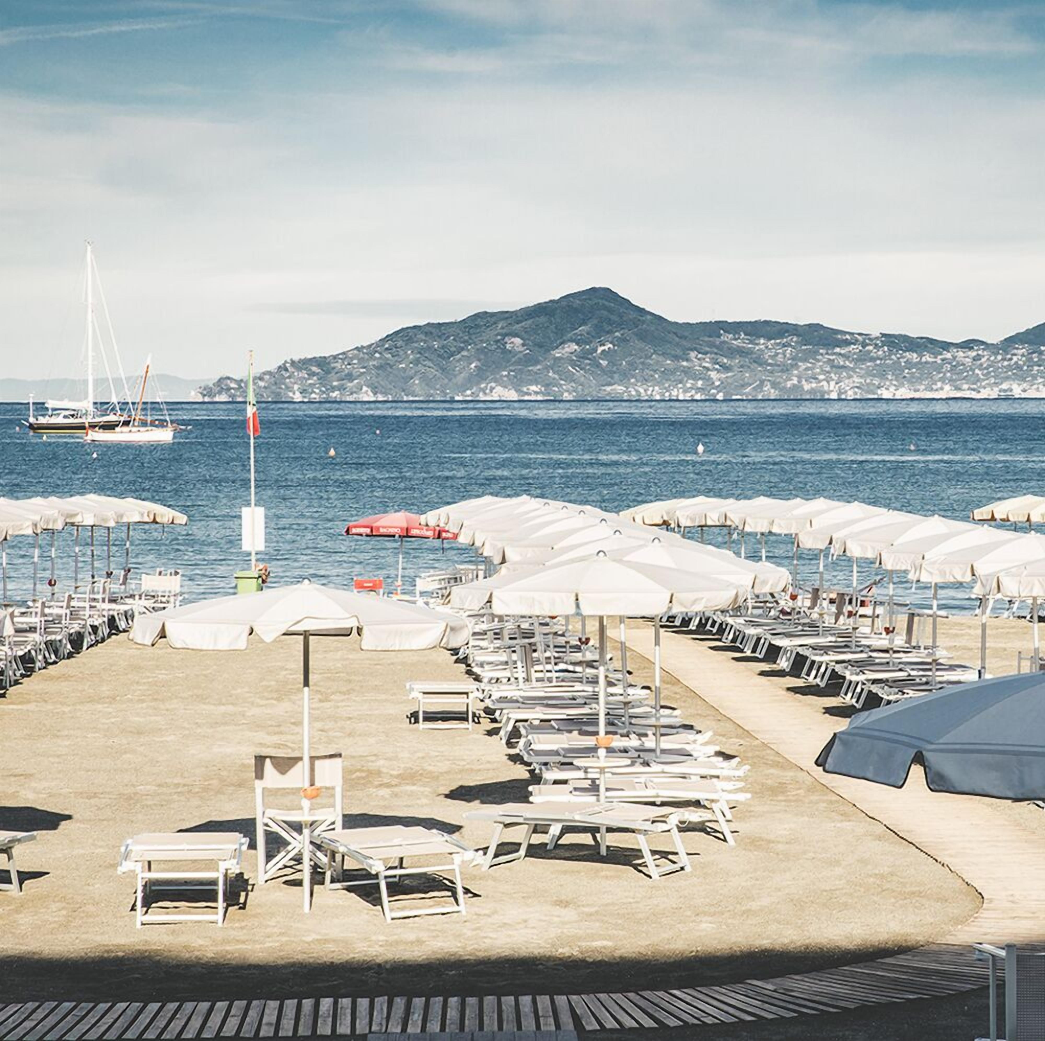 Une zone de plage bordée de rangées de chaises longues et de parasols, un voilier sur l’eau et une chaîne de montagnes en arrière-plan.