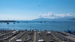 A beachside hotel with rows of sunbeds and umbrellas, a clear view of the sea, and boats in the background.