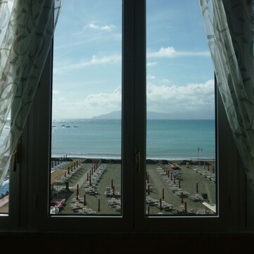 A view through a window with patterned curtains, overlooking a beach with a parking area and a distant mountain.