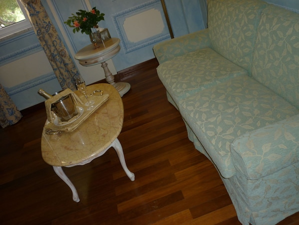Una habitación con un sofá estampado, una pequeña mesa redonda con una bandeja que sostiene una botella de champán y copas, un florero con flores y una pared decorada.