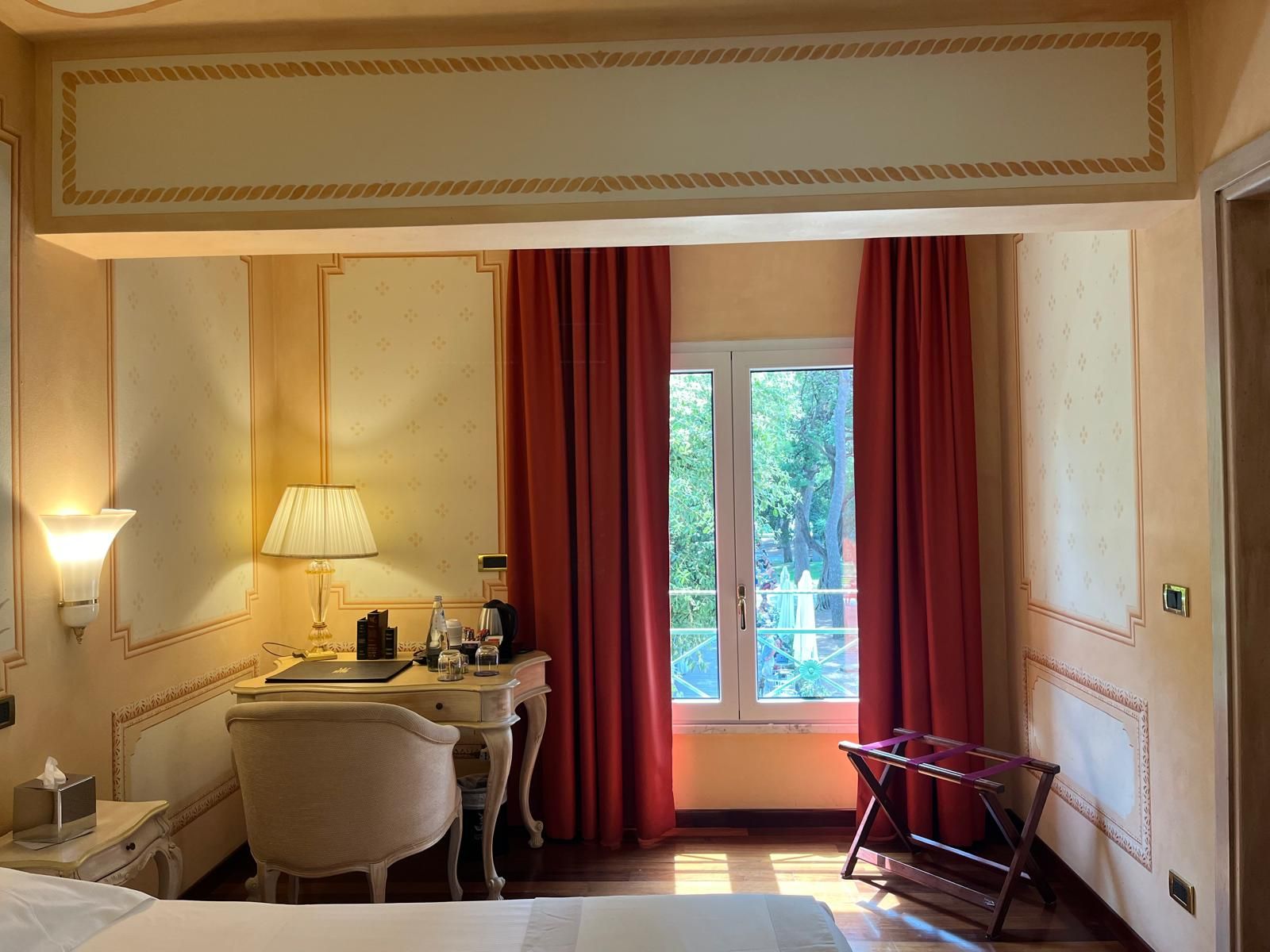 Ein Hotelzimmer mit Bett, Schreibtisch mit Lampe, Stuhl, Fenster mit roten Vorhängen und Blick ins Grüne.