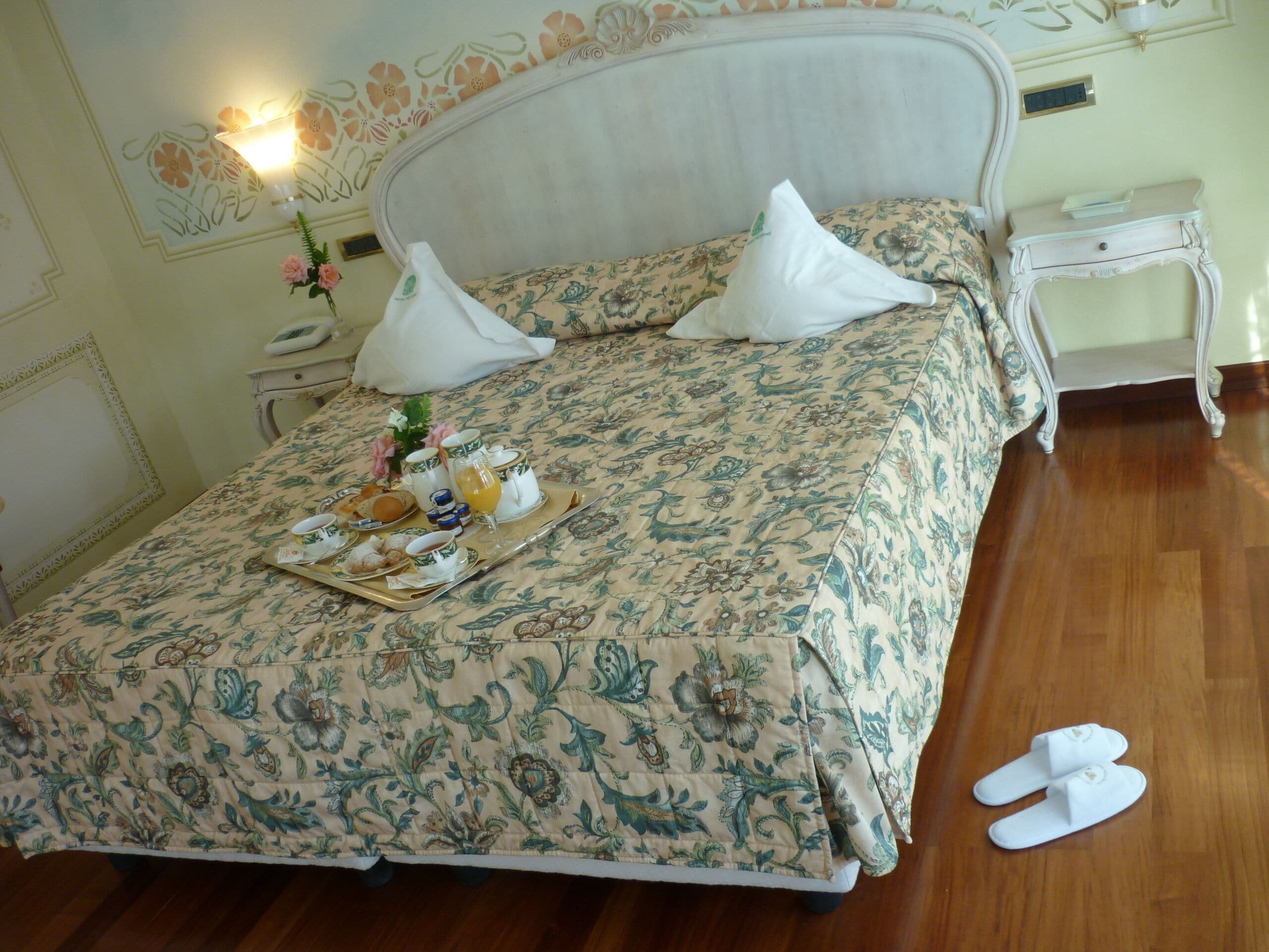 Una cama bien hecha con ropa de cama floral, una bandeja con alimentos para el desayuno y un par de pantuflas en el suelo.