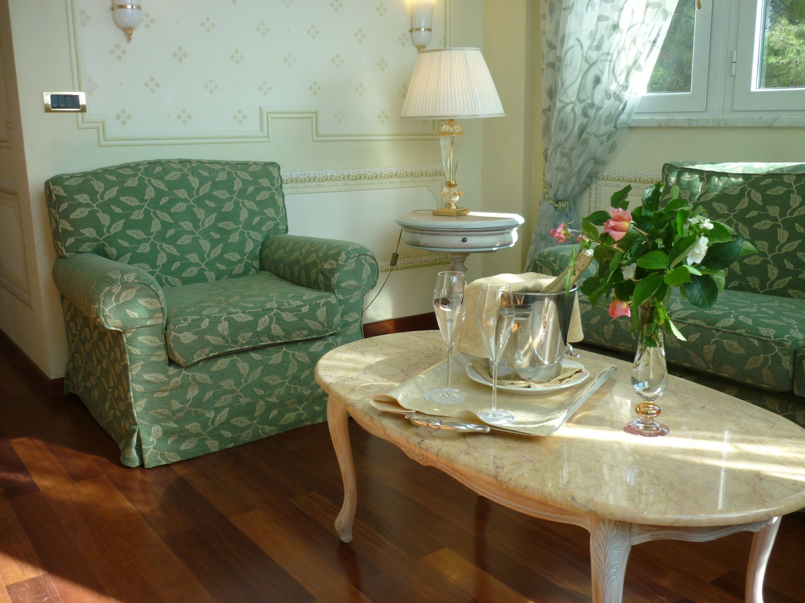 A room with a green patterned sofa, a marble table with a vase of flowers, and a lamp.