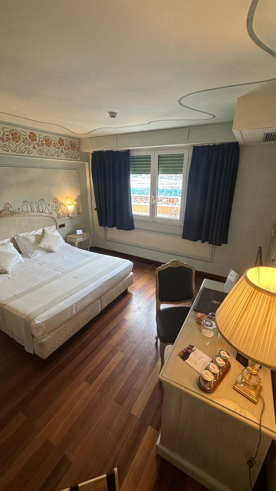 A hotel room with a bed, a desk with a lamp, a chair, a window with curtains, and a decorative wall panel.