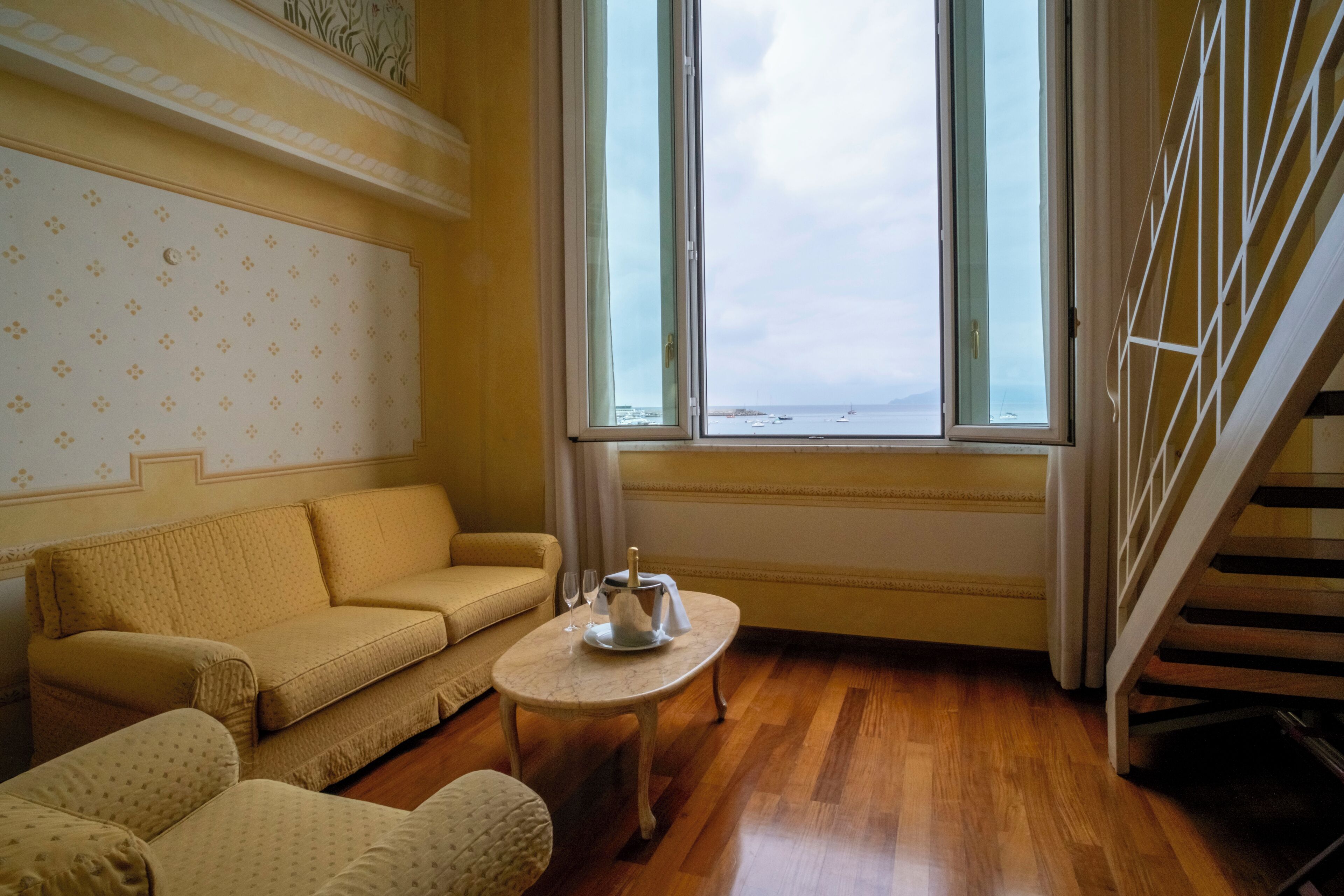 Quarto com sofá, uma mesa pequena com uma garrafa e vista para o mar através de janelas amplas.
