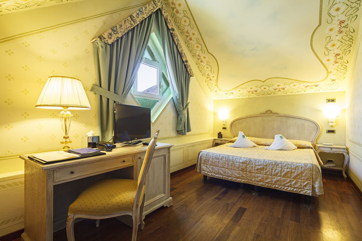 Habitación de hotel con escritorio, cama, silla, televisor y ventana con cortinas.