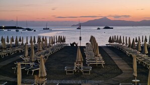 A beach with rows of sun loungers and umbrellas, boats in the water, and a mountain in the background.
