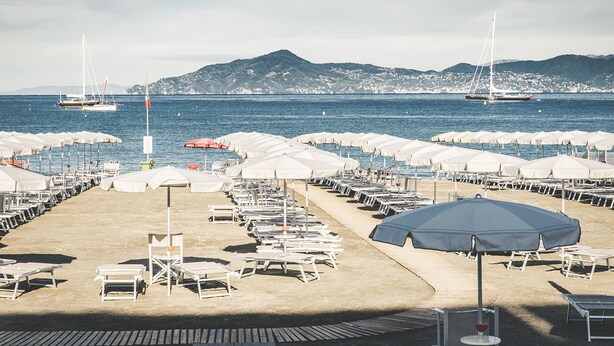 A beach with rows of sun loungers and umbrellas, boats in the water, and mountains in the background.