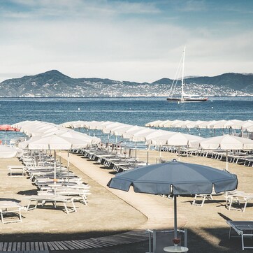 A beach with rows of sun loungers and umbrellas, boats in the water, and mountains in the background.