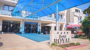 A hotel entrance with a blue canopy, a sign reading "Hotel Royal", and a building with balconies and a shop in the background. - Hotel Royal (Golden Sands)