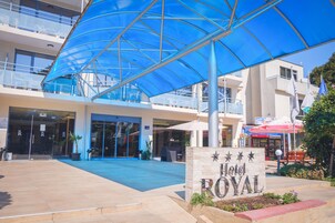 Entrada de un hotel con un toldo azul, un letrero que dice "Hotel Royal" y un edificio con balcones y una tienda de fondo.
