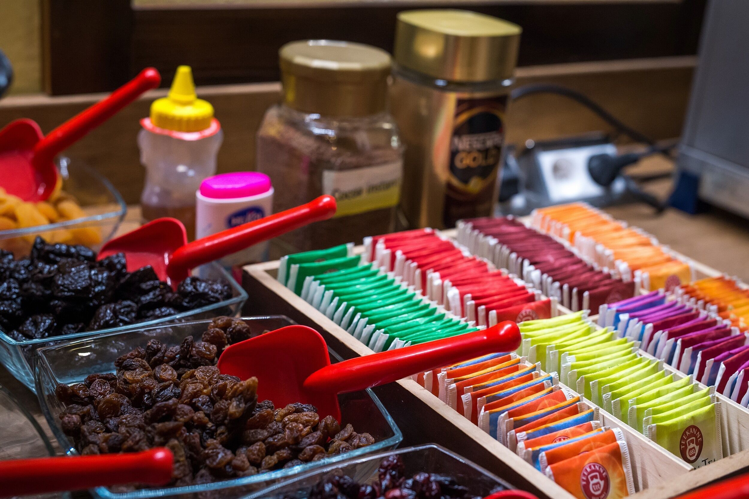 A variety of food items, including dried fruits and packaged snacks, arranged in clear containers with red scoops.
