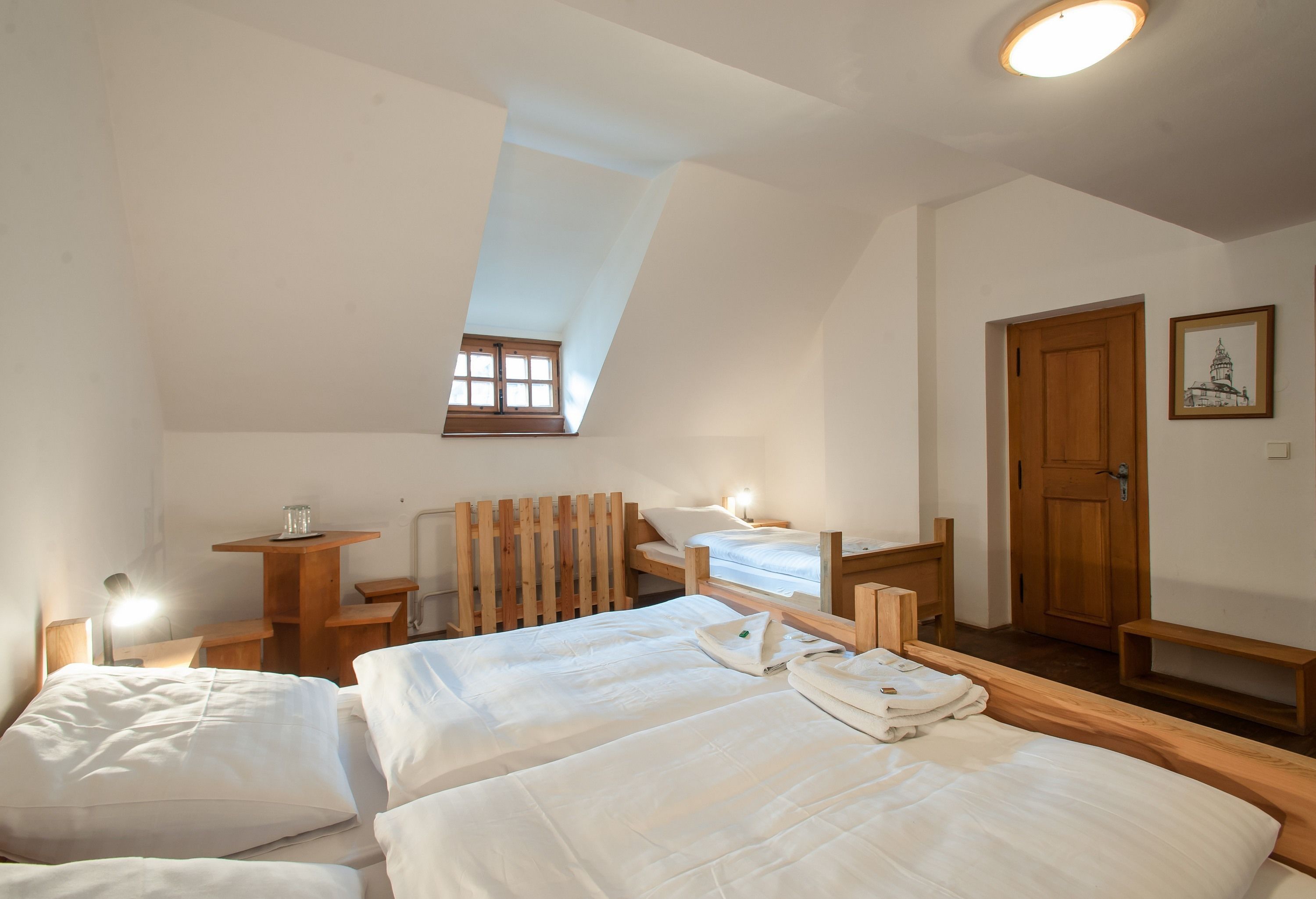 A room with two beds, a wooden desk, a small table, a window, and a door.