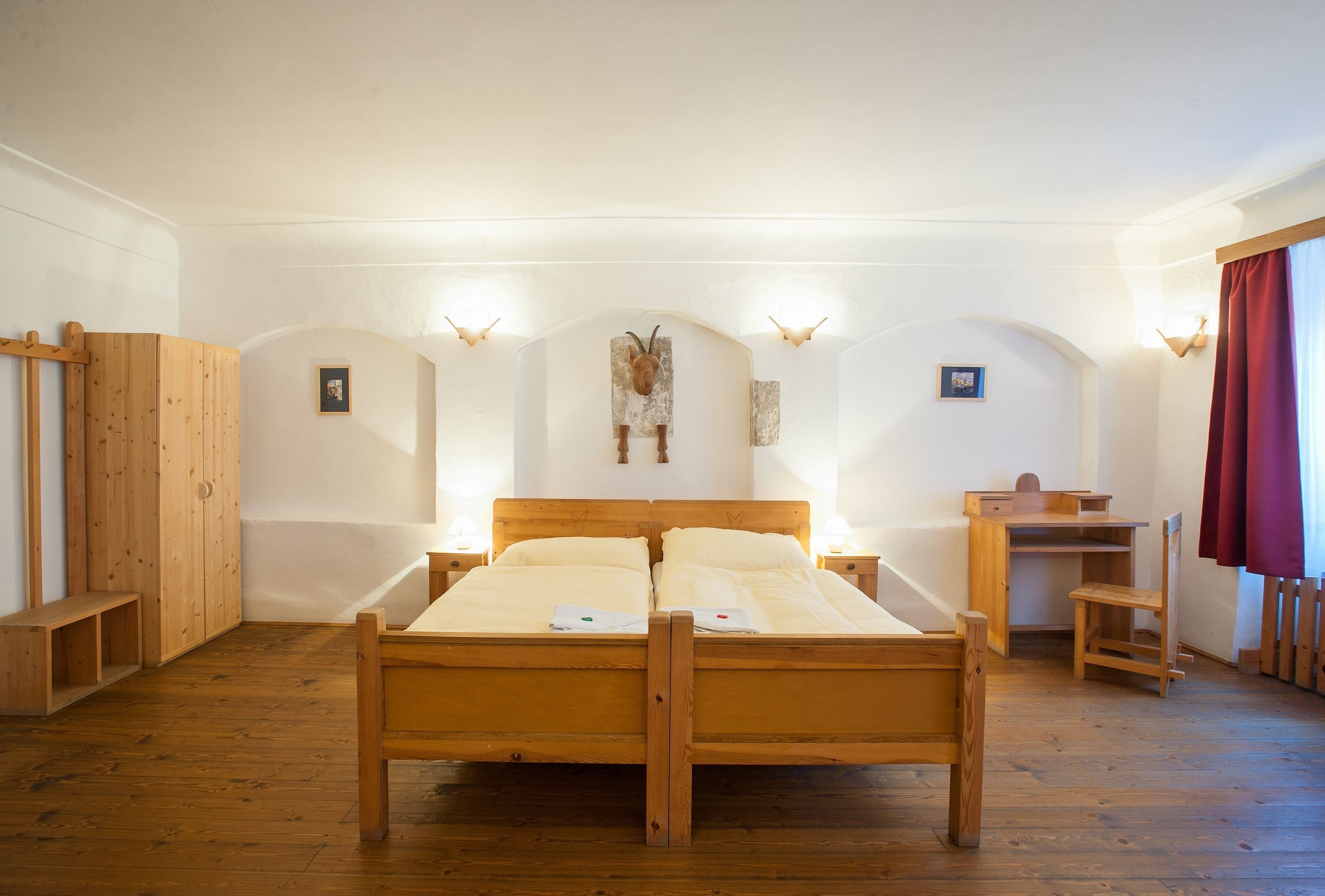 A wooden bed with two pillows, a wooden desk, and a bench in a room with white walls and wooden flooring.