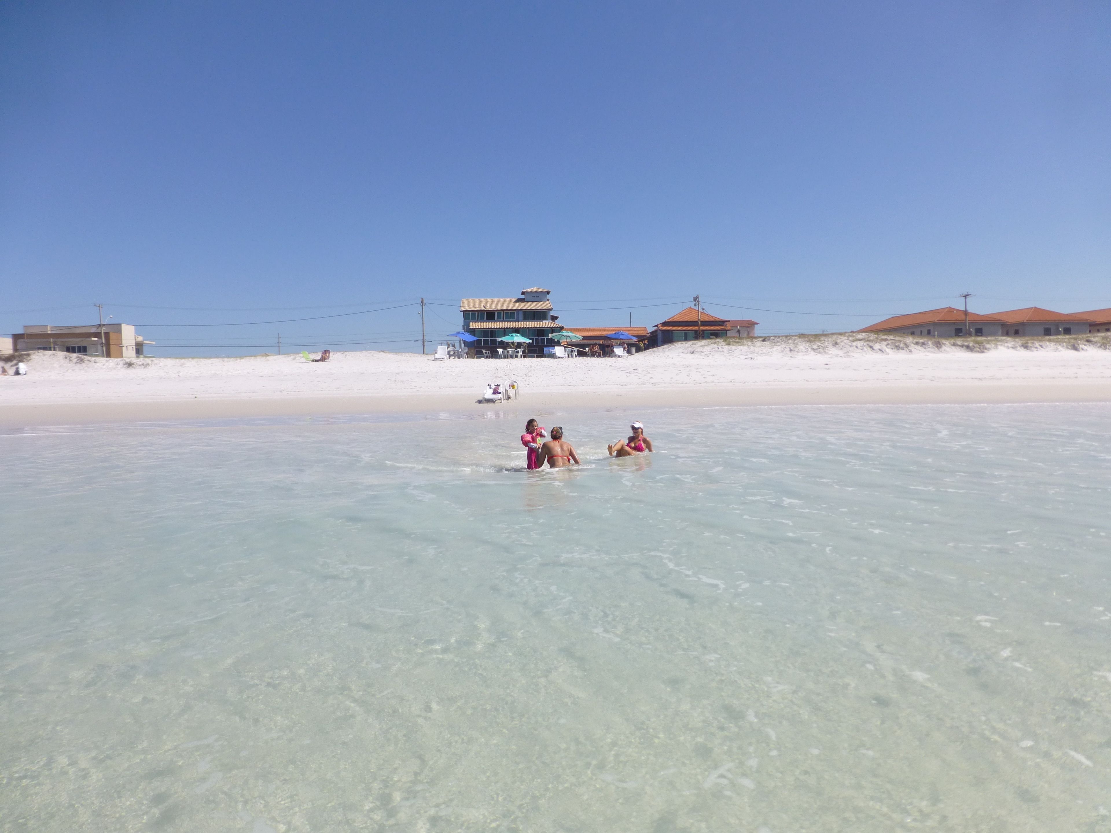 A beach with clear water, a few people in the water, and buildings in the background.