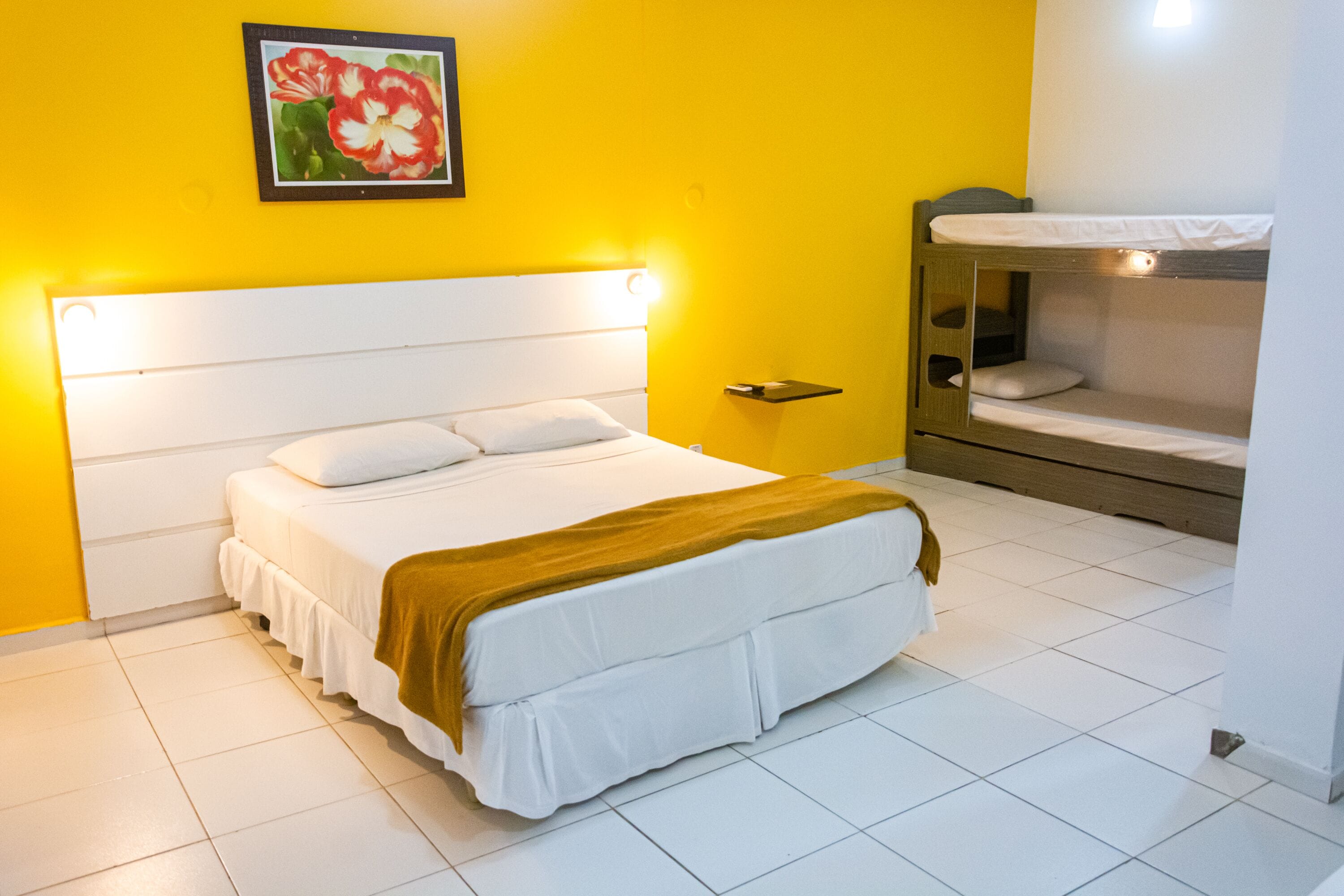 Quarto de hotel com cama, mesa de cabeceira, um banco e luminária fixada na parede.