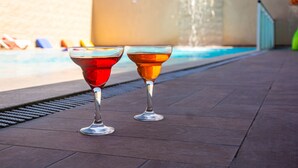 Two glasses of red and amber beverages on a tiled surface near a pool with a water feature. - Pousada Laguna (Cabo Frio)