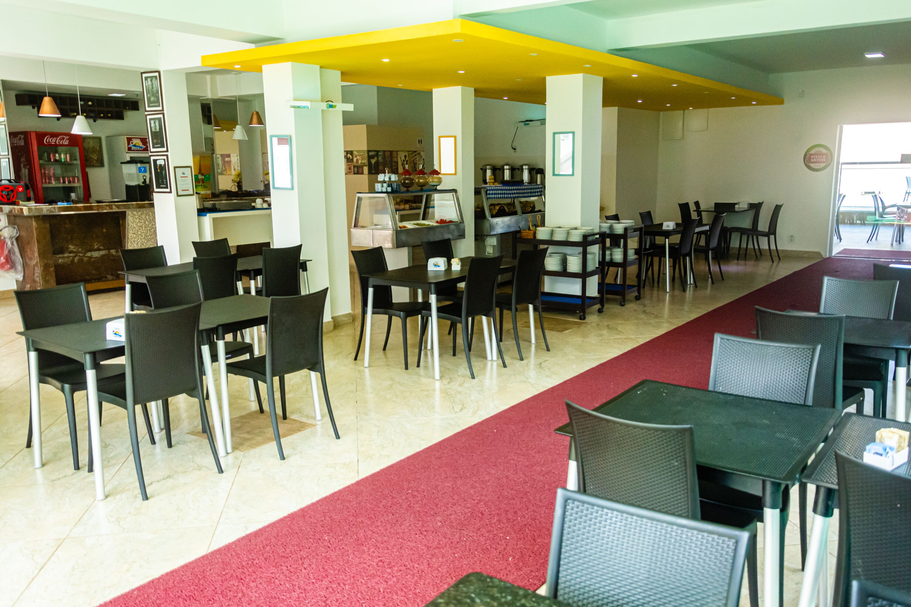 A restaurant with multiple tables and chairs, a red carpet, and a Coca-Cola refrigerator.