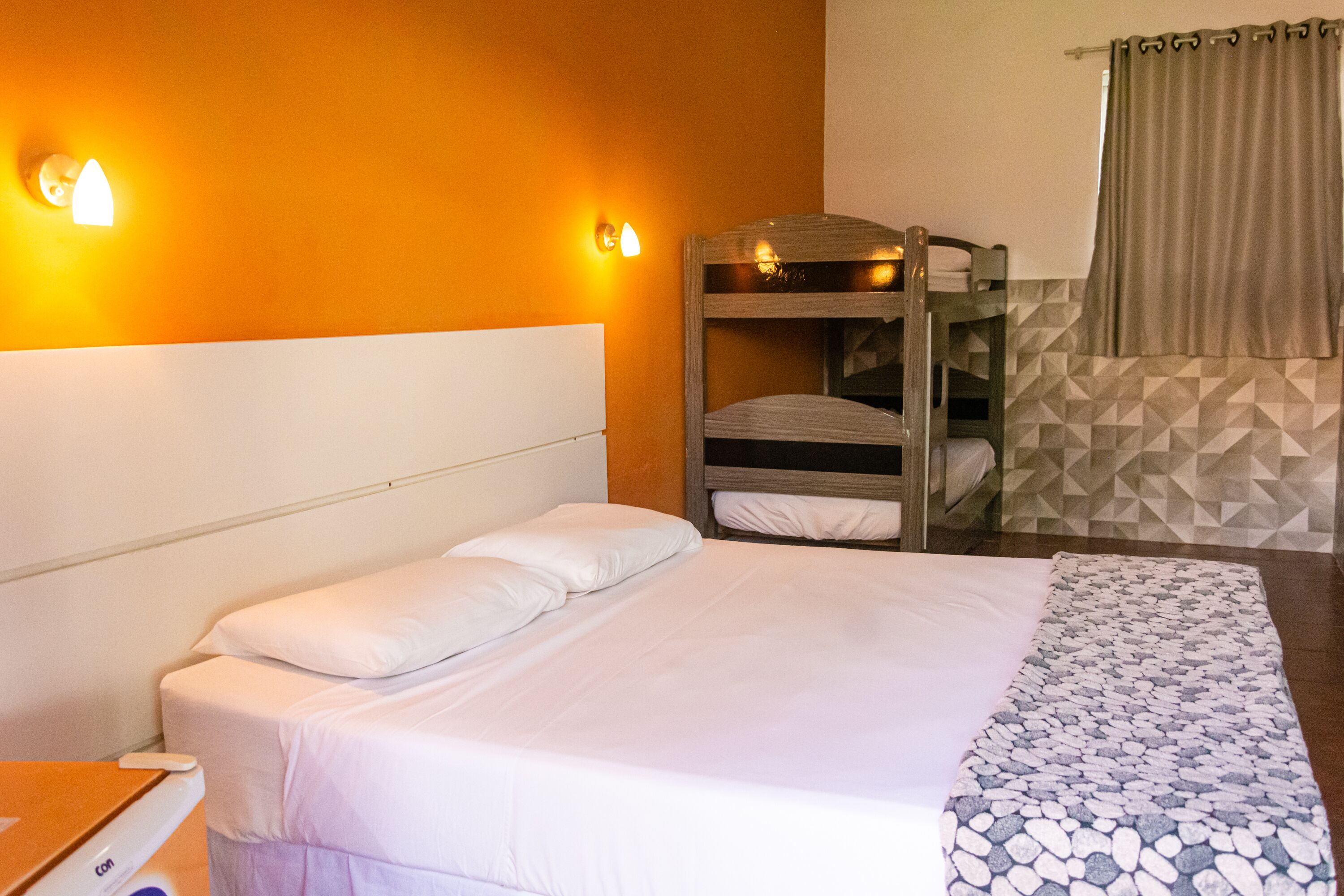 A hotel room with a bunk bed, a single bed with white linens, a patterned throw blanket, and a wall-mounted light fixture.