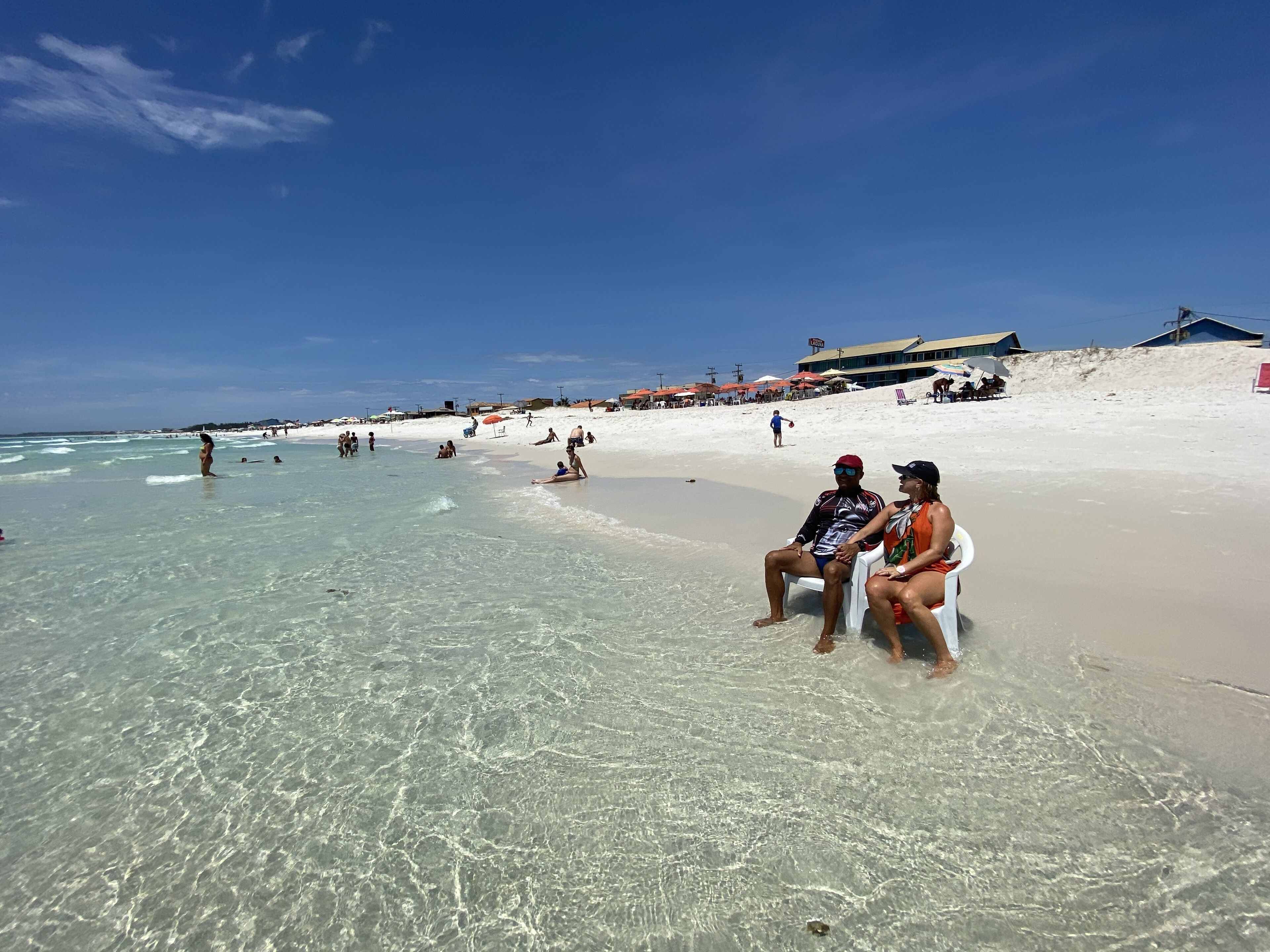 Een strandzicht met helder ondiep water, mensen die op het zand ontspannen, en enkele gebouwen op de achtergrond.