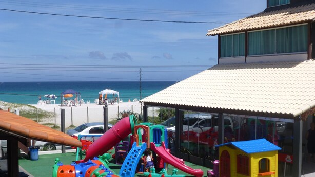 A playground with slides and climbing structures, a beach in the background, and a building with a tiled roof. - Pousada Laguna (Cabo Frio)
