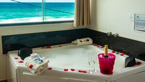 A room with a large window overlooking the ocean, a bathtub with rose petals and champagne, and a white towel on the edge. - Pousada Laguna (Cabo Frio)