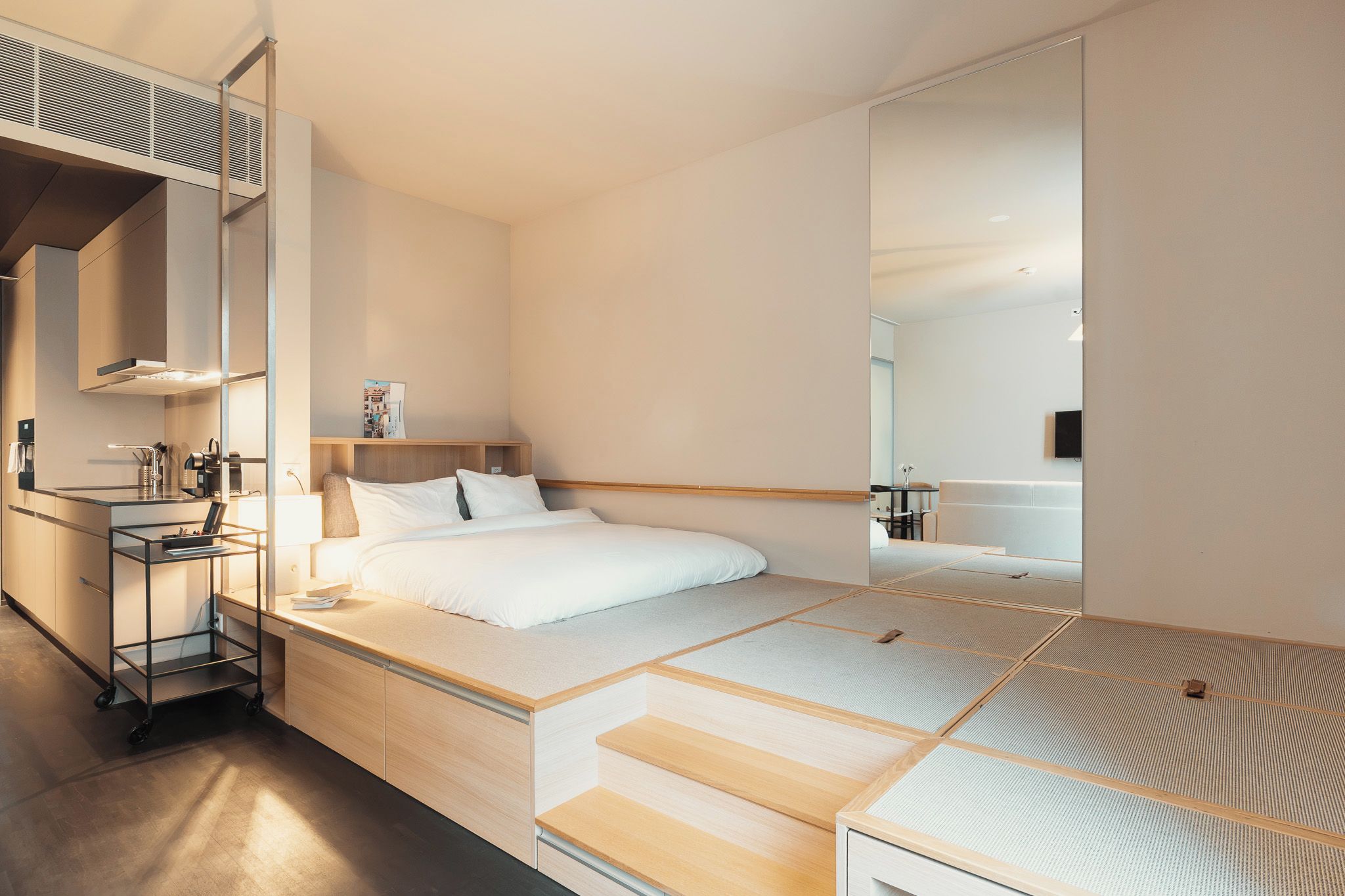 A modern hotel room with a bed, a bedside table, a mirror, and a kitchenette area.
