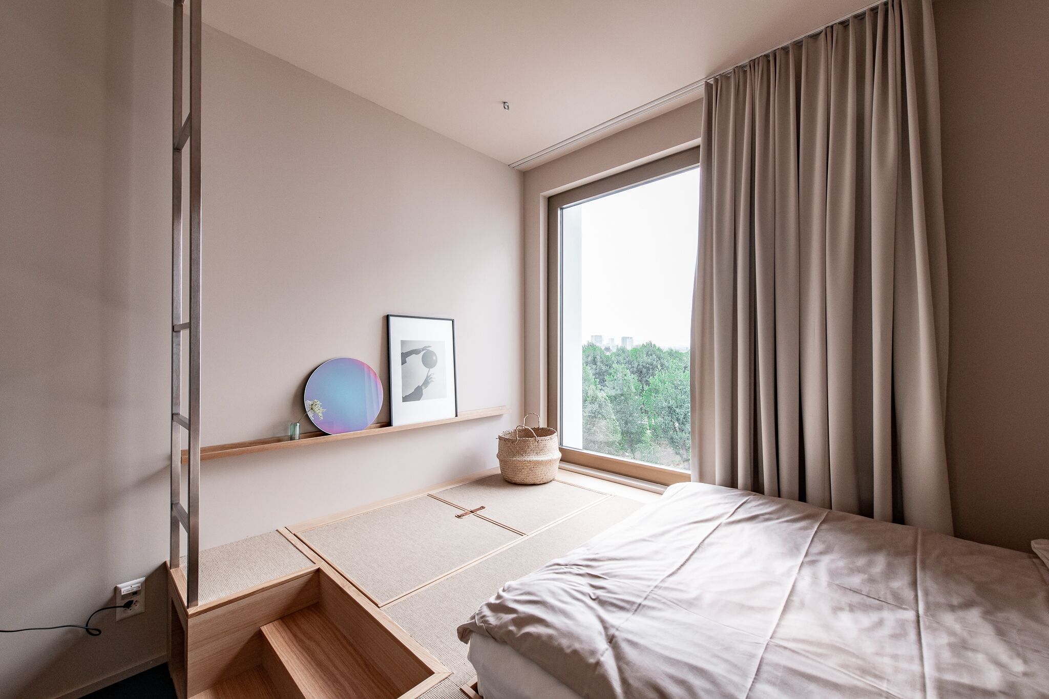 Une chambre à coucher au style minimaliste, avec un lit, une fenêtre avec des rideaux, une étagère avec des photos encadrées et une petite table en bois.