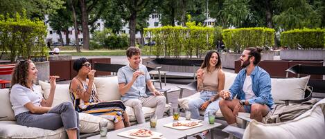 A group of people sitting on outdoor furniture in a park-like setting with trees and a building in the background.